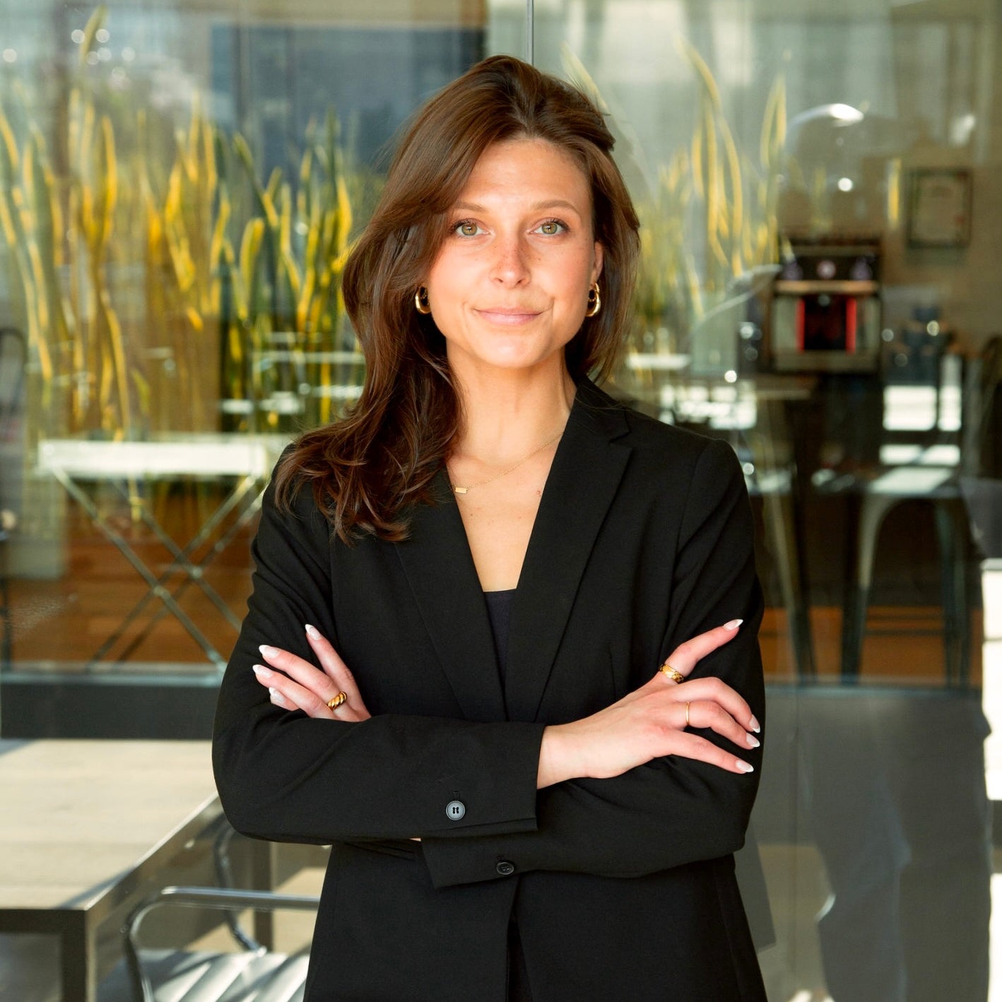 Professional portrait of a woman standing indoors in a modern office setting. She is wearing a black blazer and black top, with her arms crossed and a slight smile. Her shoulder-length brown hair is styled to one side, and she is wearing small gold hoop earrings and delicate jewelry. Behind her, glass walls reveal tall green plants, office furniture, and soft natural light, creating a polished, contemporary workplace atmosphere.