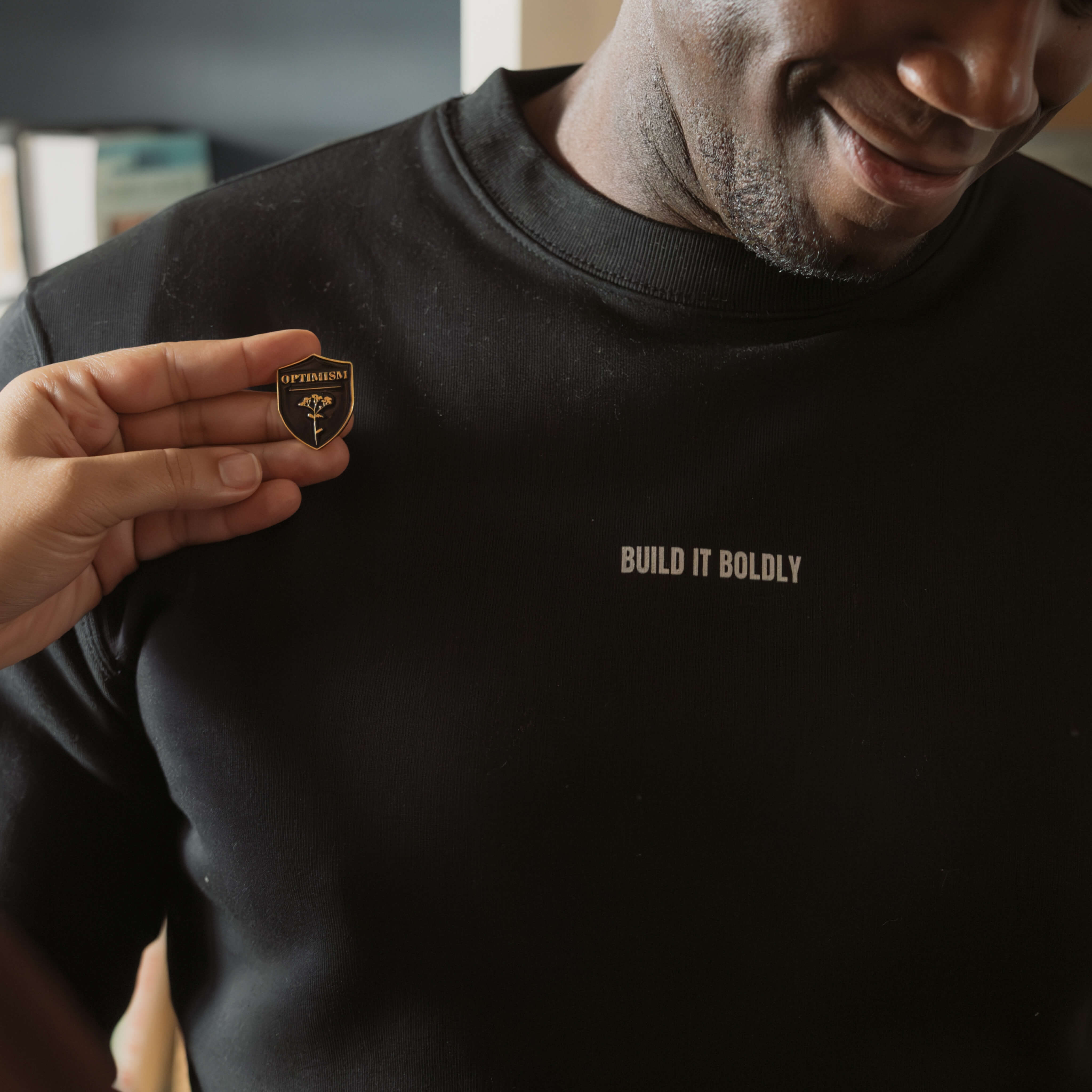 Hand holding an Optimism pin beside a man wearing a black Build It Boldly crewneck sweatshirt with white chest logo.