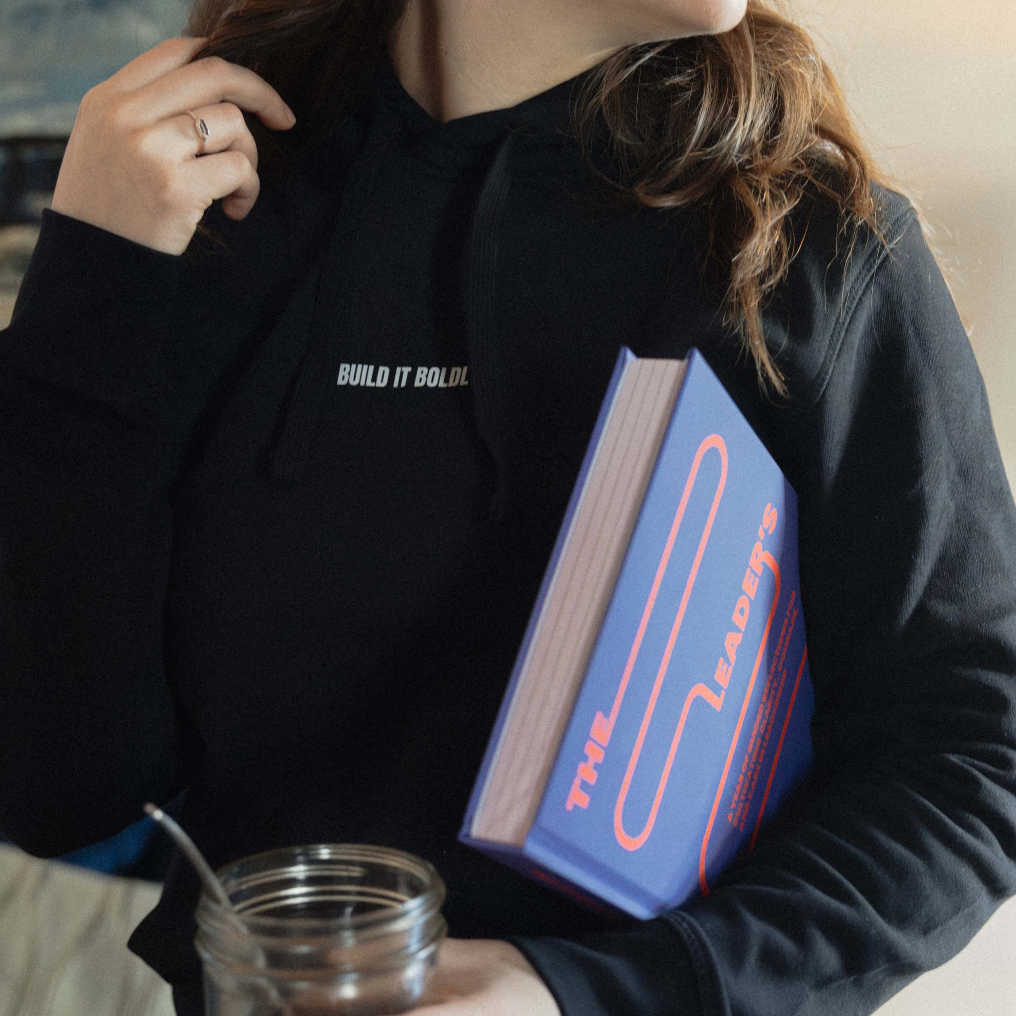 lose-up of a woman wearing a black Build It Boldly hoodie and holding The Leader’s Work book.