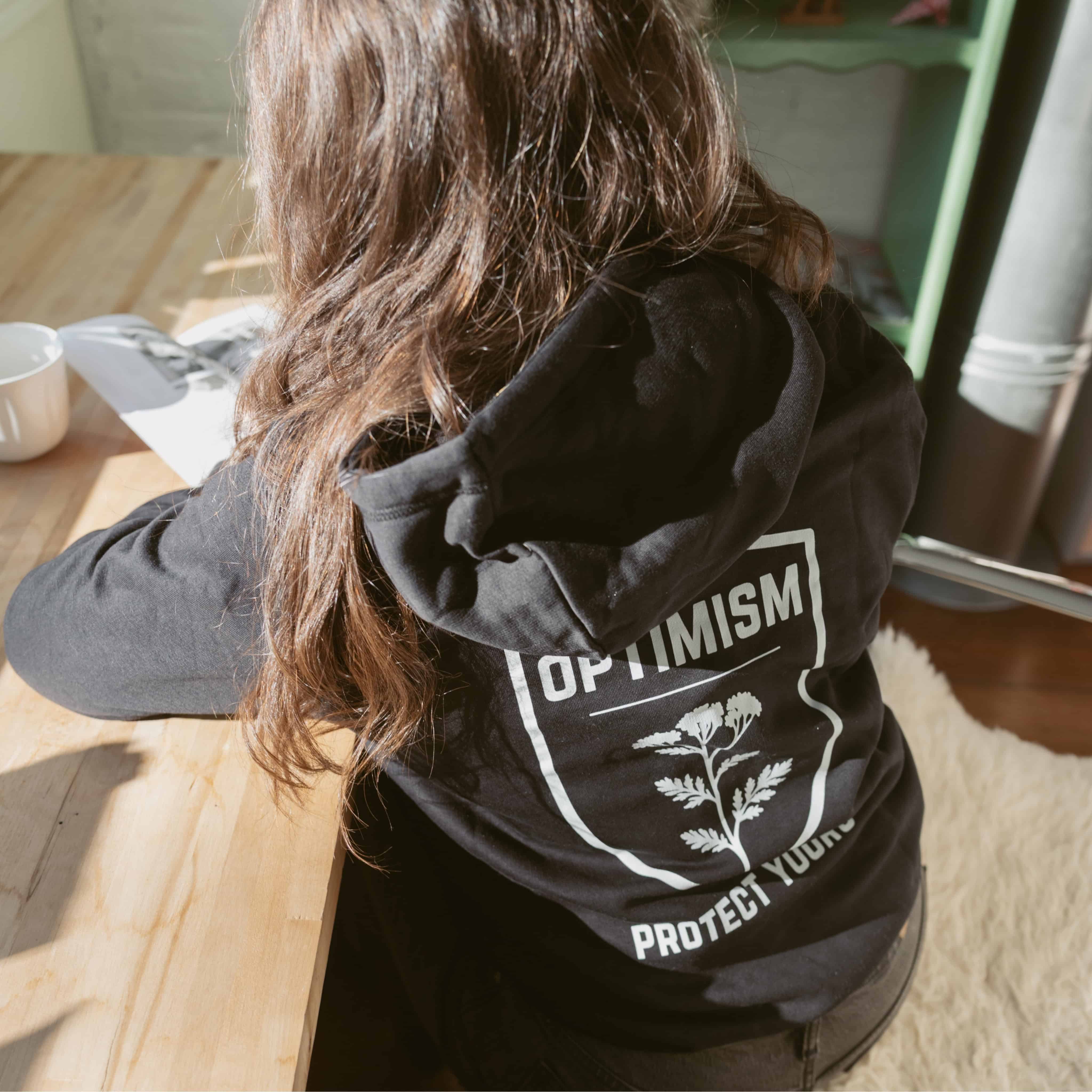 Woman seated at a desk wearing a black Build It Boldly hoodie with large “Optimism Protect Yours” graphic on the back.