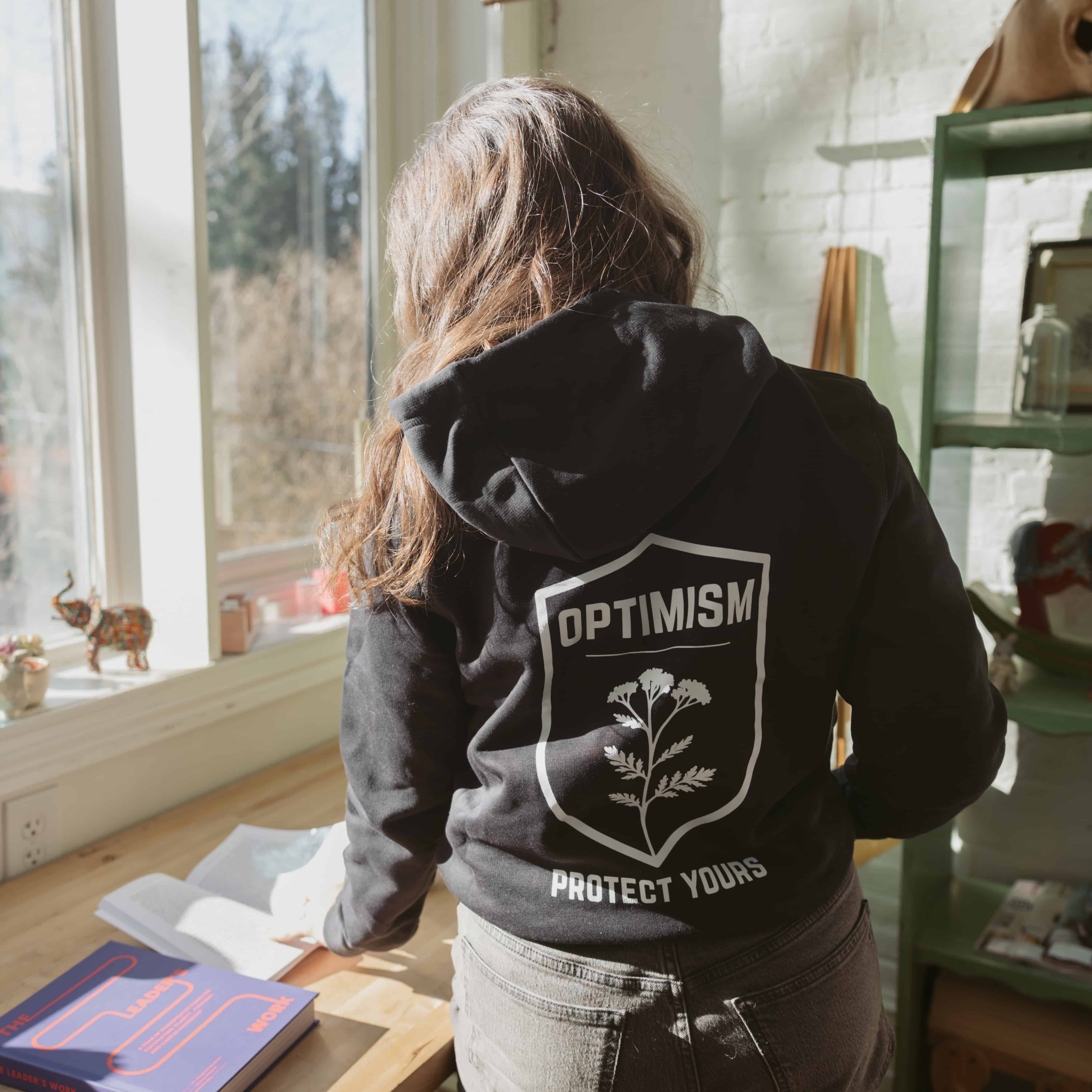 Woman standing by a window in a black Build It Boldly hoodie with “Optimism Protect Yours” back graphic in a sunlit studio.