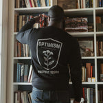 Back view of a man wearing a black Build It Boldly sweatshirt with large “Optimism Protect Yours” graphic while placing a book on a bookshelf.