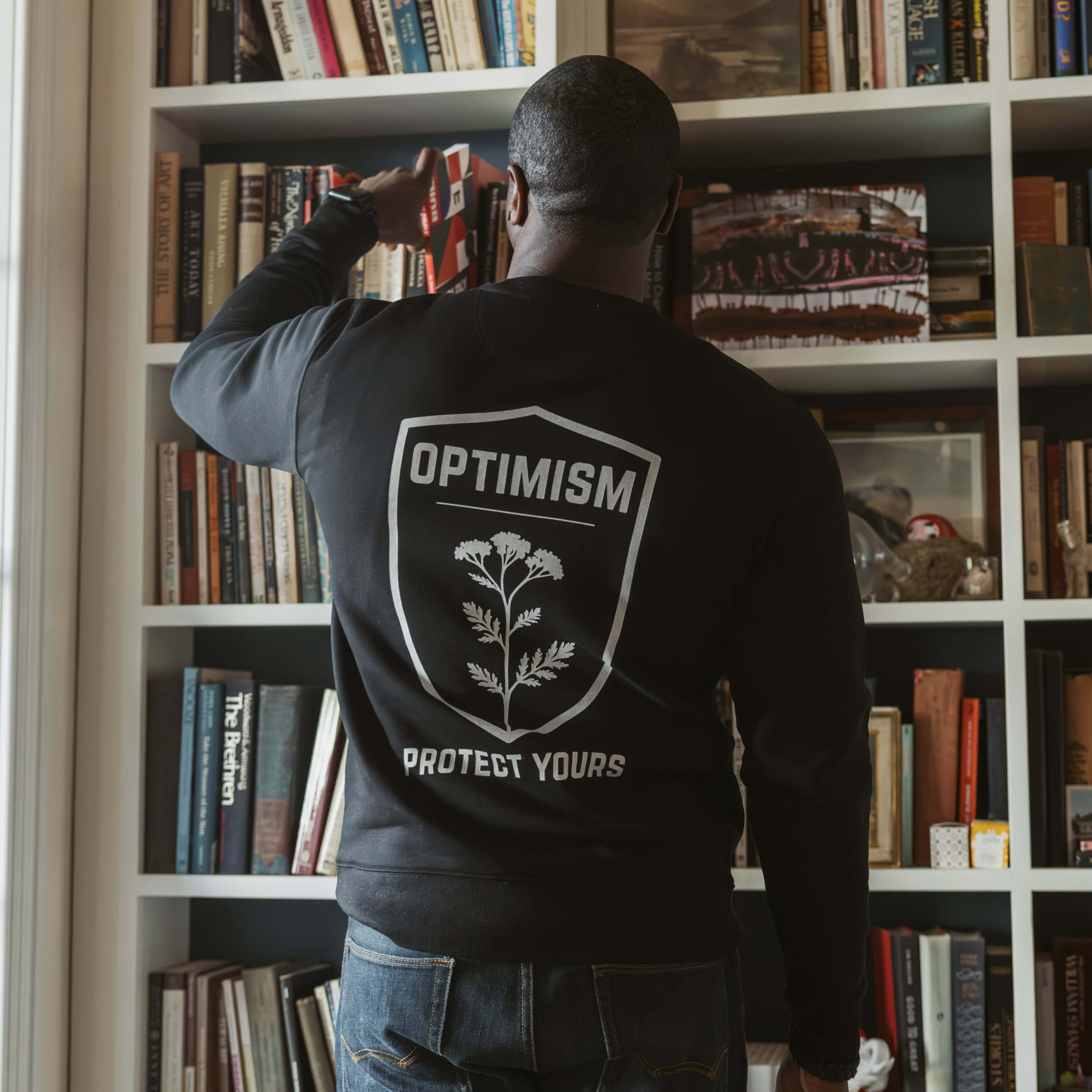 Back view of a man wearing a black Build It Boldly sweatshirt with large “Optimism Protect Yours” graphic while placing a book on a bookshelf.