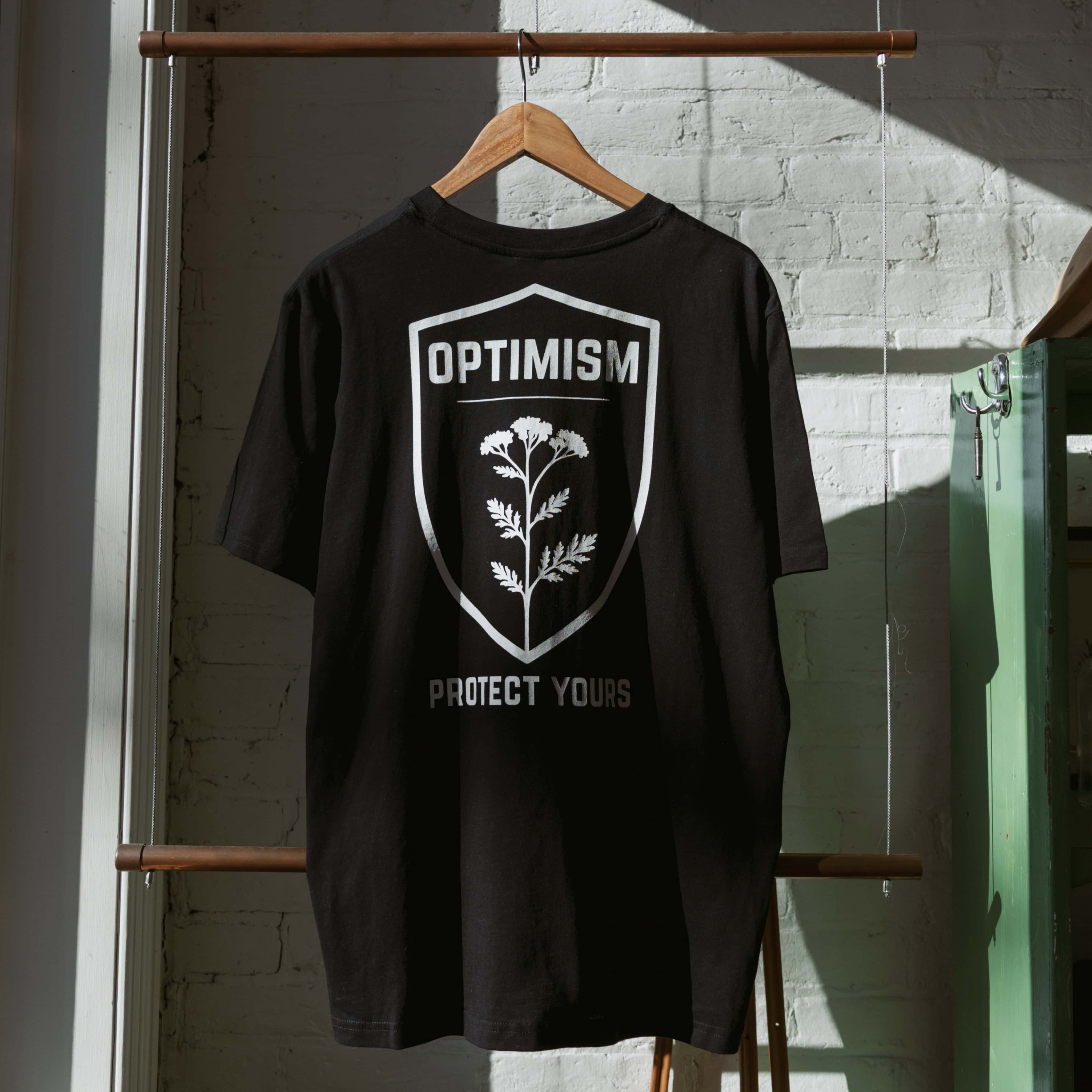 Black Build It Boldly t-shirt hanging in sunlight, showing the back “Optimism Protect Yours” shield graphic on a wooden hanger.