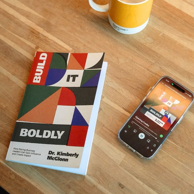 Build It Boldly book by Dr. Kimberly McGlonn next to smartphone playing audiobook and coffee mug on wooden table