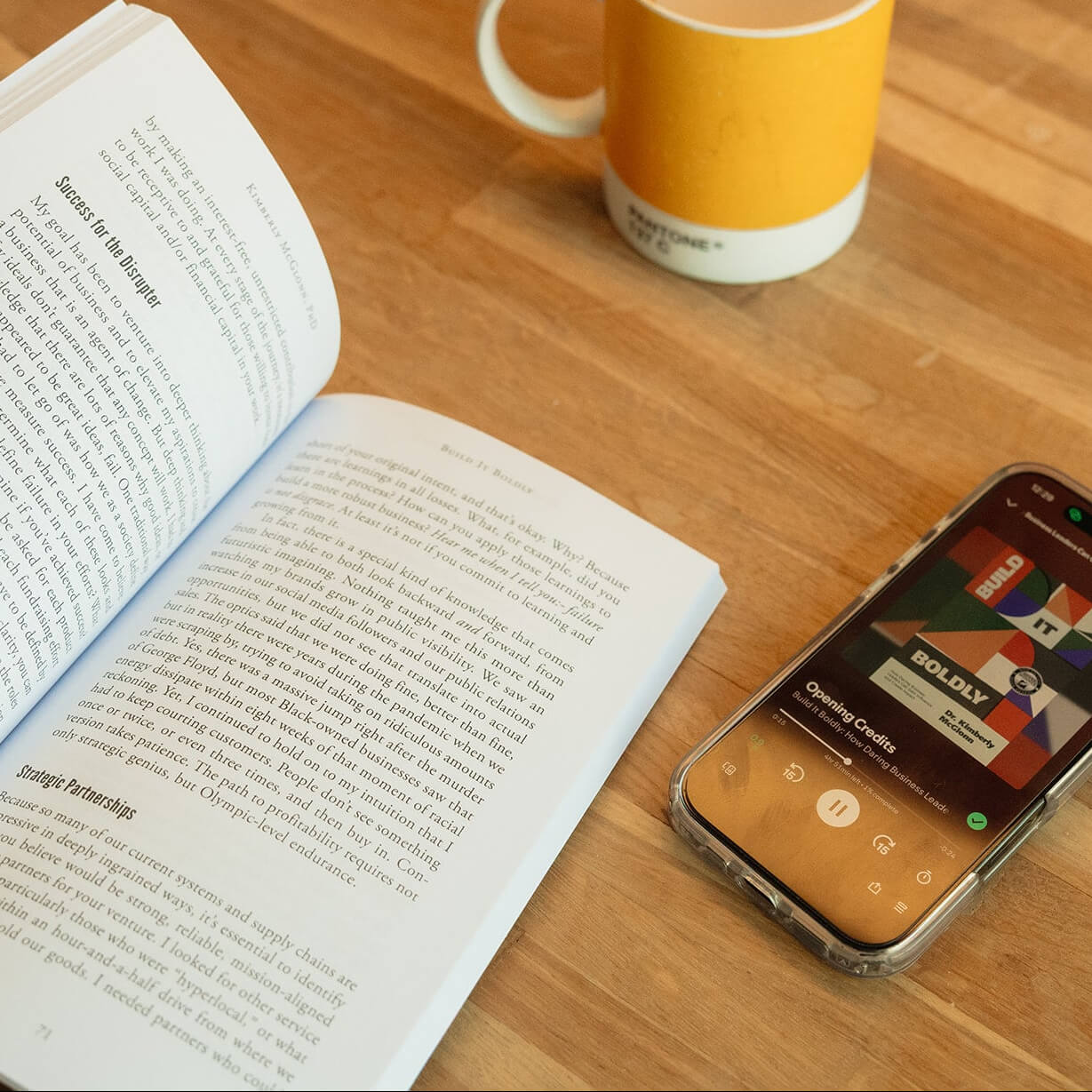 Open copy of Build It Boldly on a table beside a phone playing the audiobook and a coffee mug.