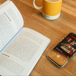 Open copy of Build It Boldly on a table beside a phone playing the audiobook and a coffee mug.