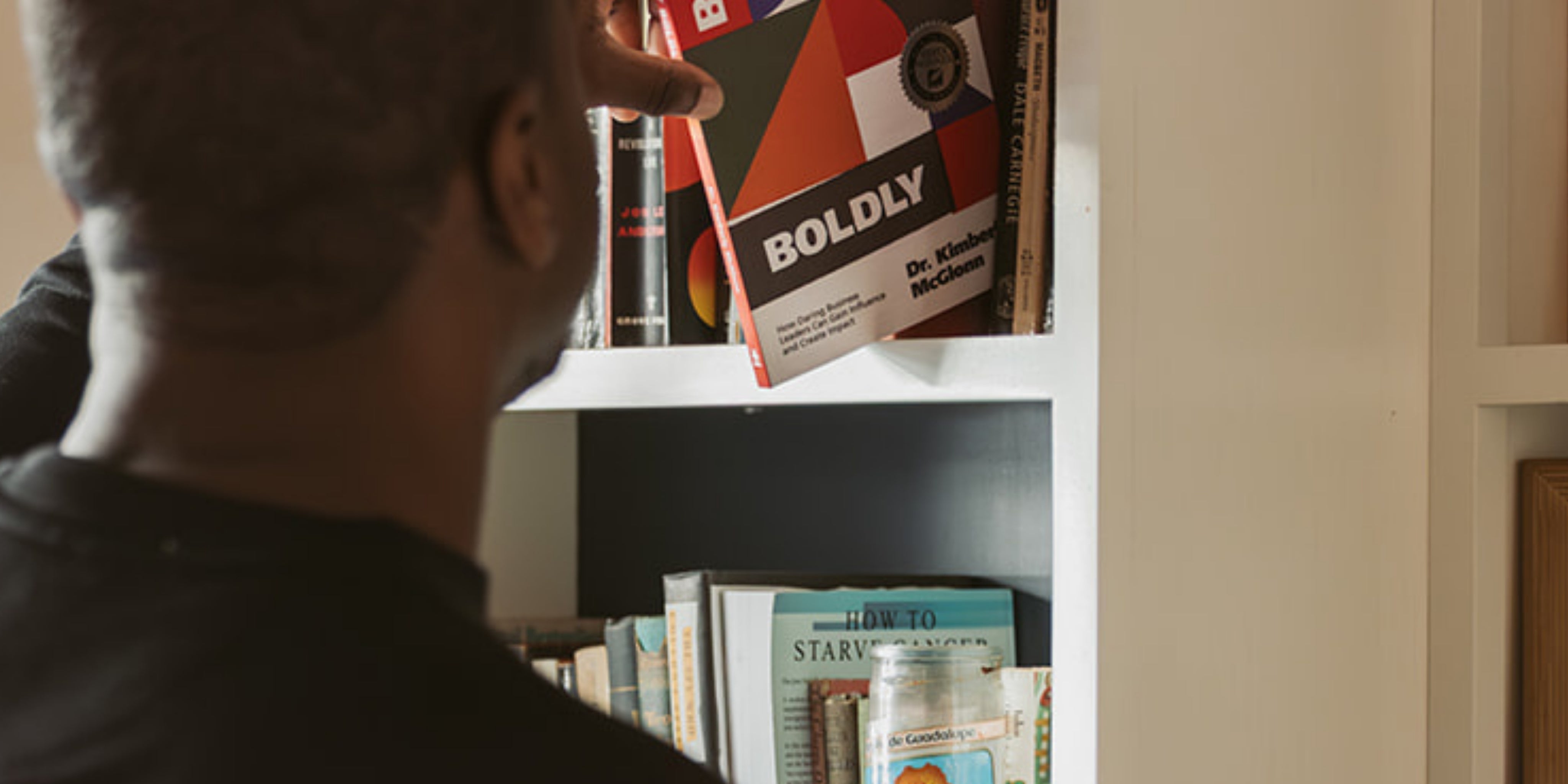 Person placing a copy of Build It Boldly by Dr. Kimberly McGlonn onto a bookshelf at home.