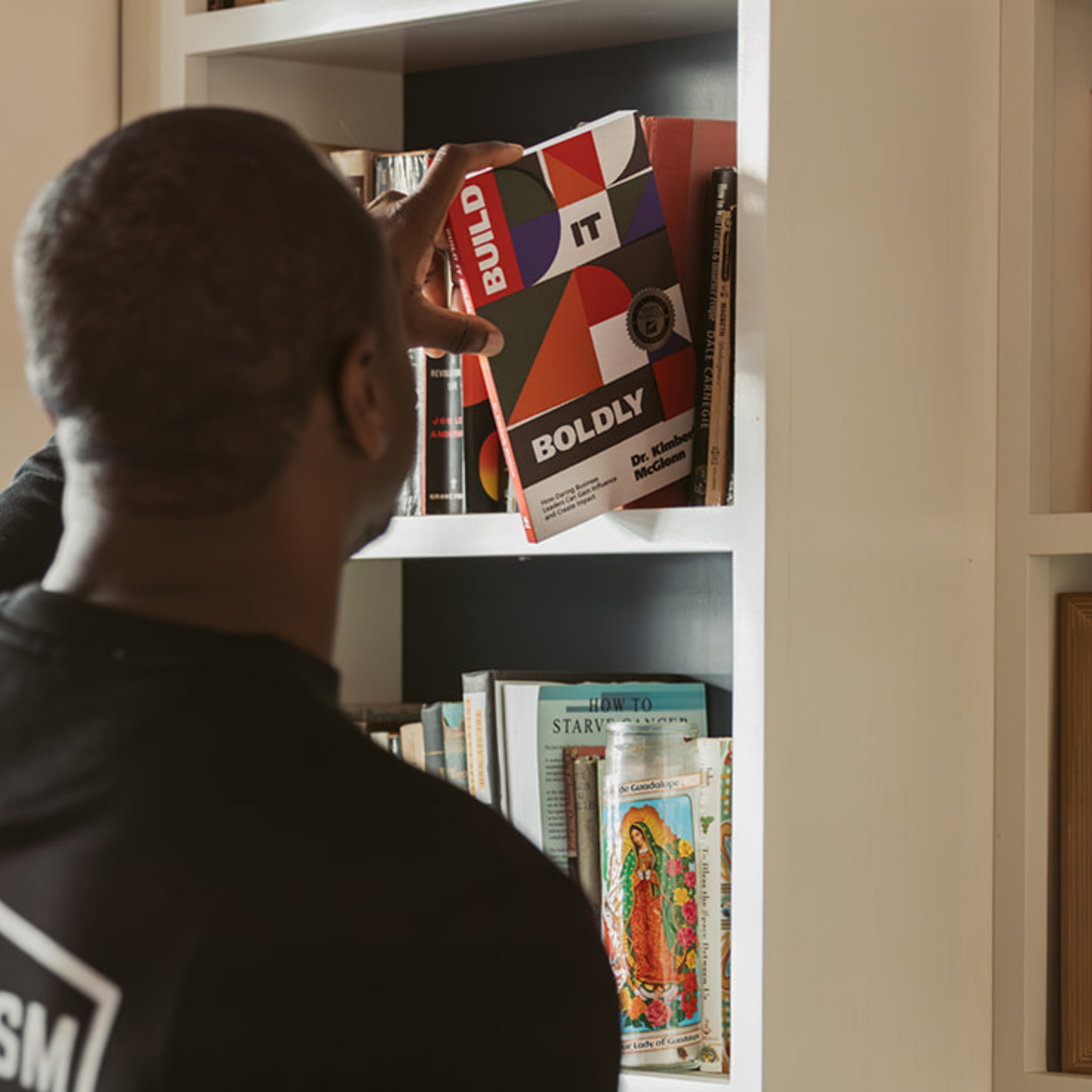 Person placing a copy of Build It Boldly by Dr. Kimberly McGlonn onto a bookshelf at home.