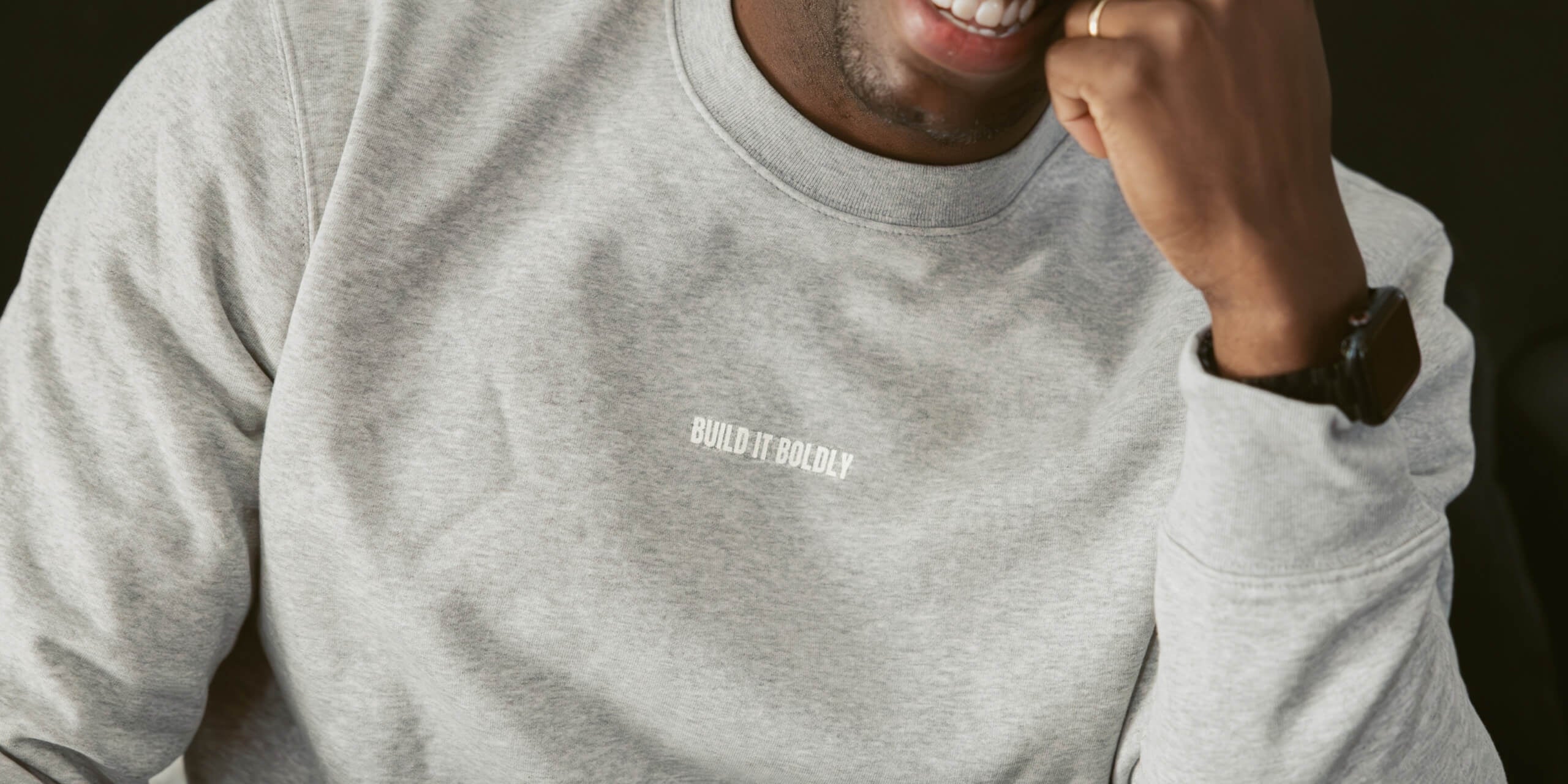 Close-up of a smiling man wearing a heather gray Build It Boldly crewneck sweatshirt with minimalist white logo on the chest.