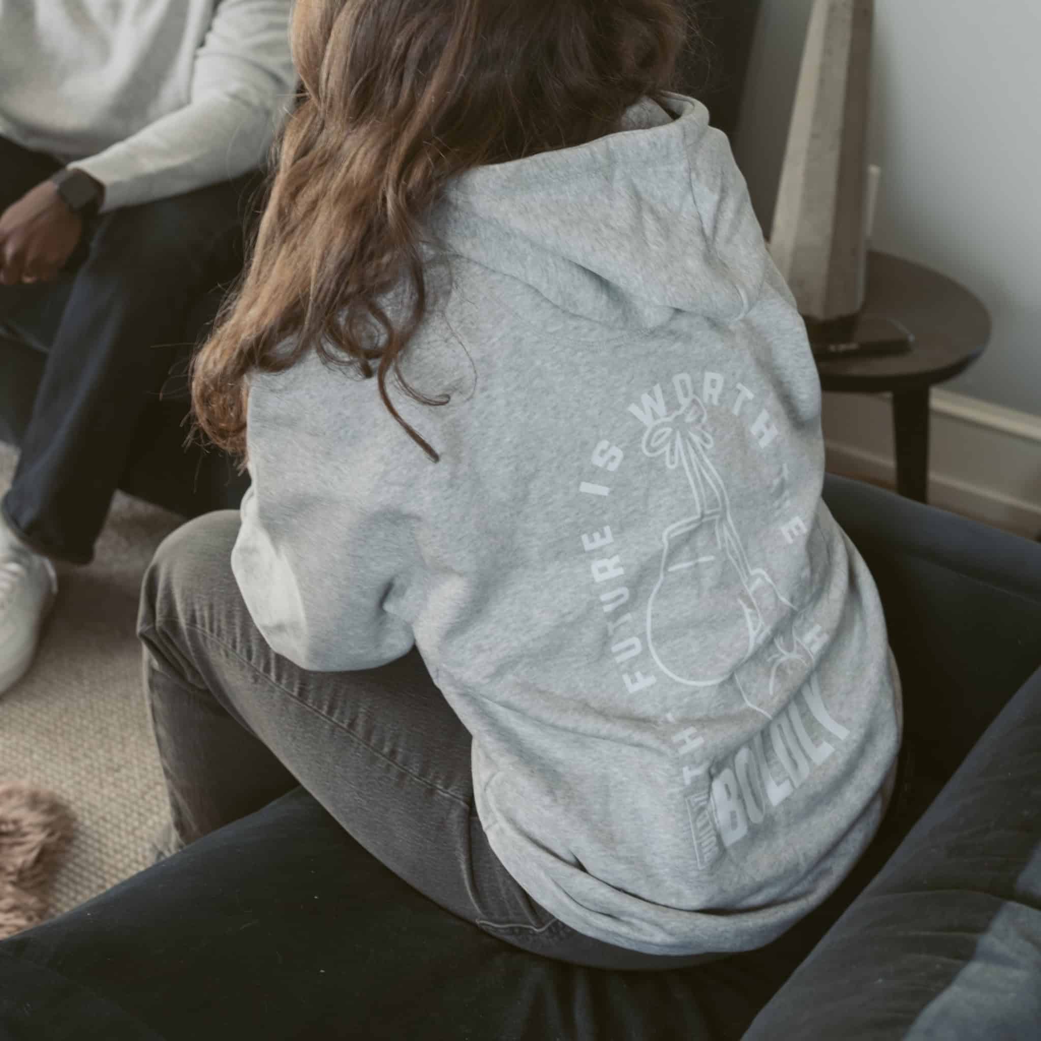 Back view of a heather gray Build It Boldly hoodie with boxing gloves graphic and “The Future Is Worth the Fight” design in a lifestyle setting.
