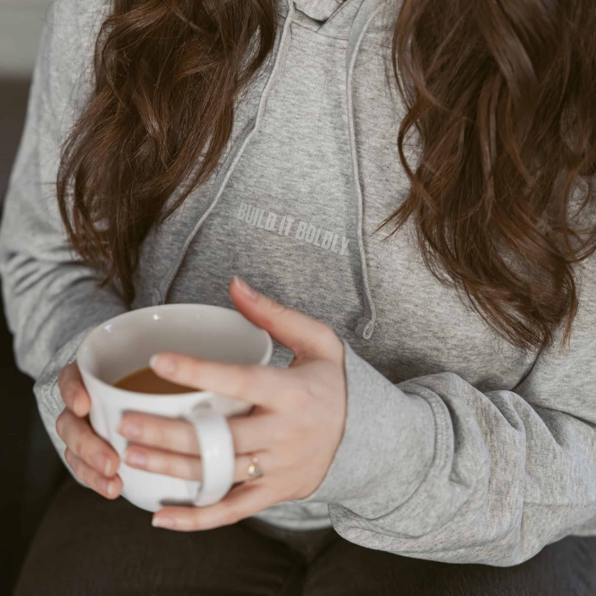Close-up of a heather gray Build It Boldly hoodie with small chest logo, styled as casual lifestyle apparel while holding a mug.