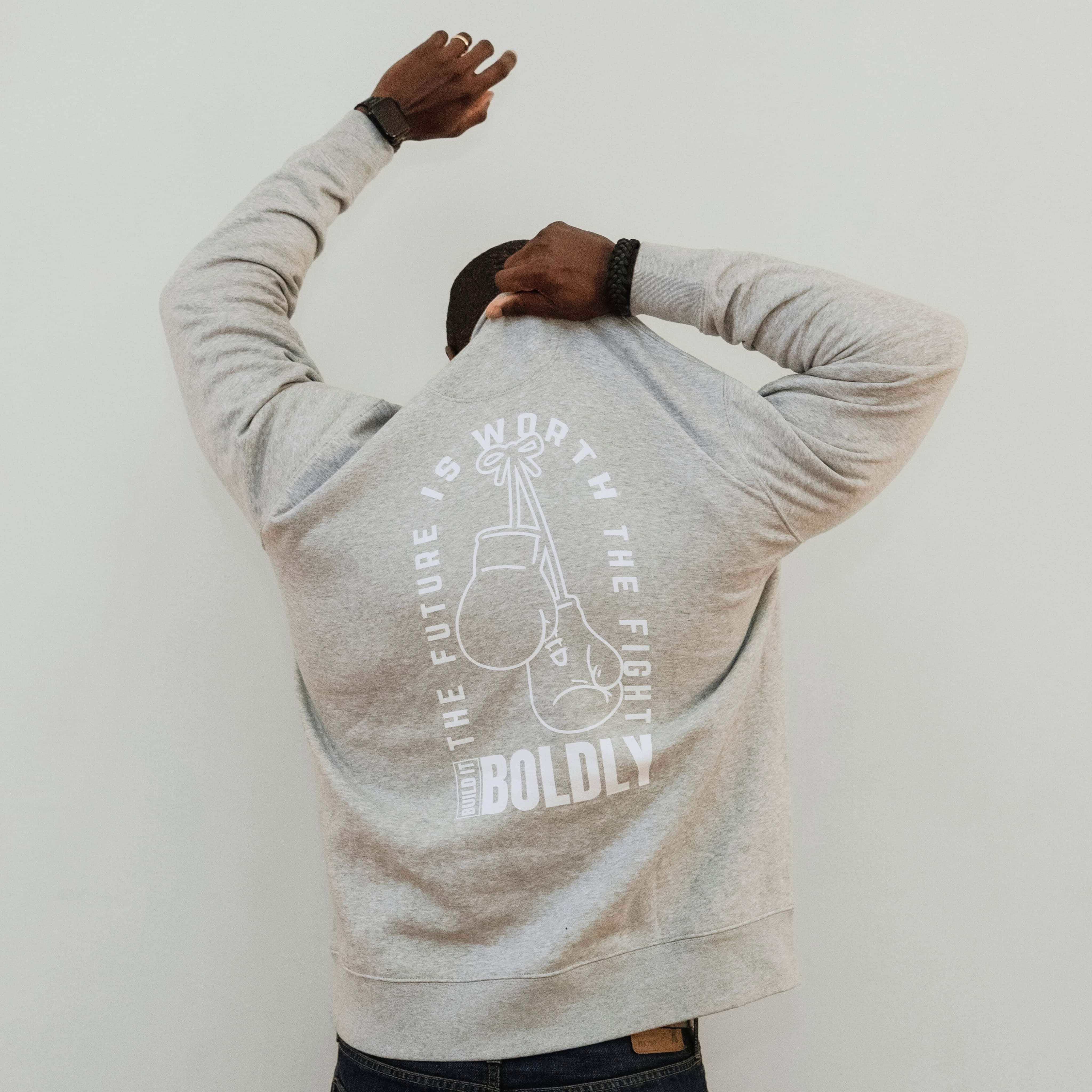 Back view of a man wearing a heather gray Build It Boldly sweatshirt with “The Future Is Worth the Fight” boxing gloves graphic.