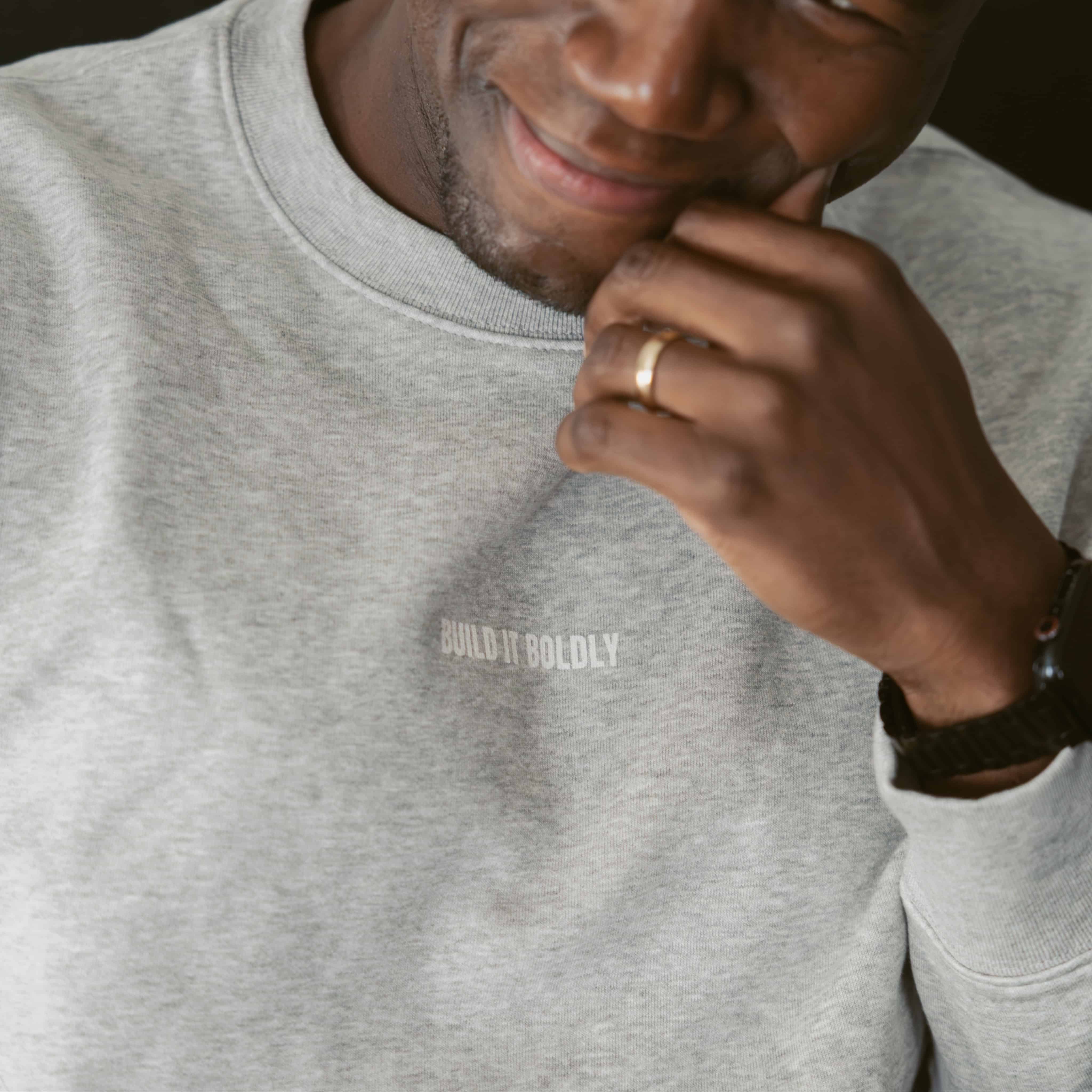Close-up of a smiling man wearing a heather gray Build It Boldly sweatshirt with small white chest logo.