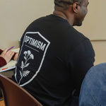 Back of a black Build It Boldly sweatshirt featuring the large “Optimism” shield graphic during a workshop