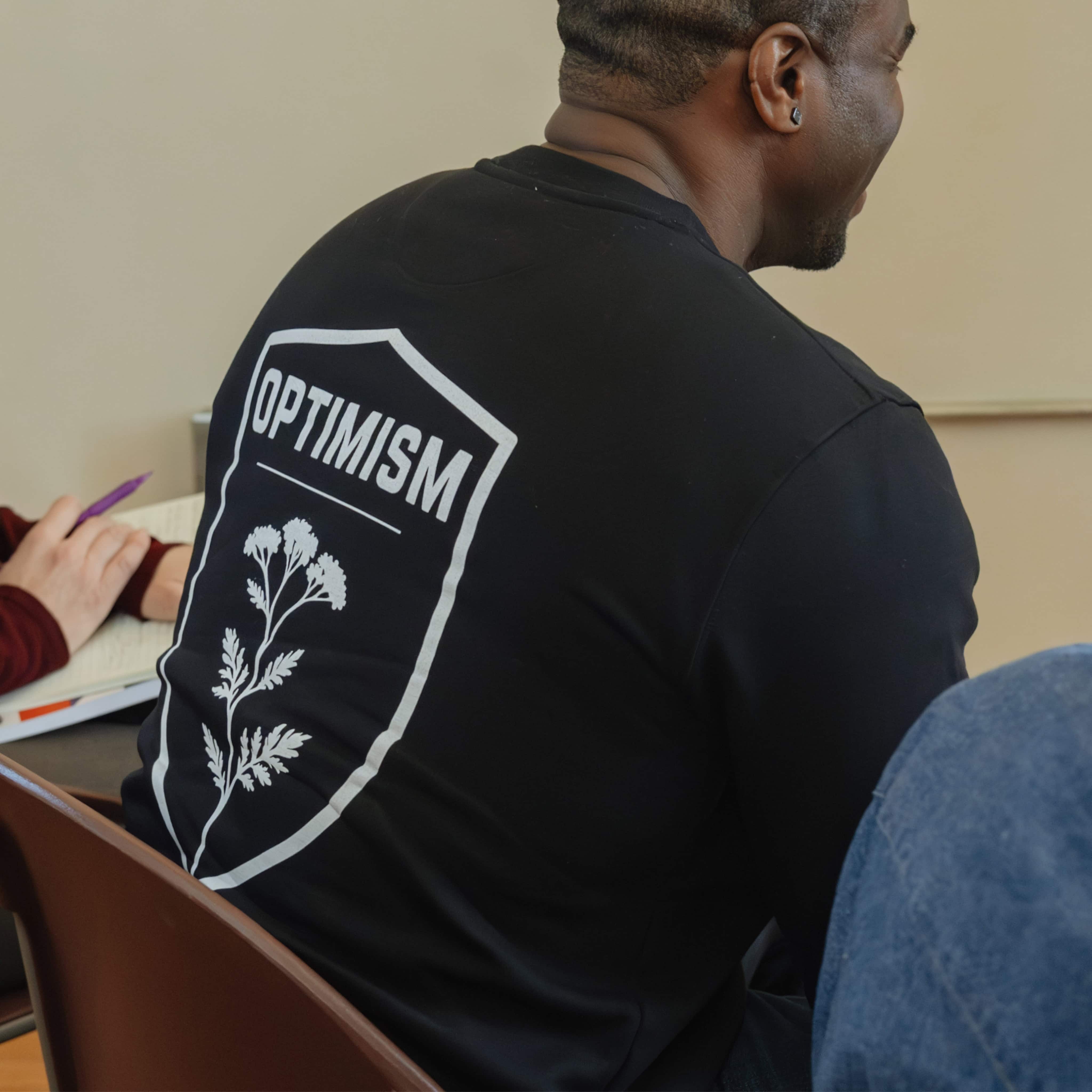 Back of a black Build It Boldly sweatshirt featuring the large “Optimism” shield graphic during a workshop