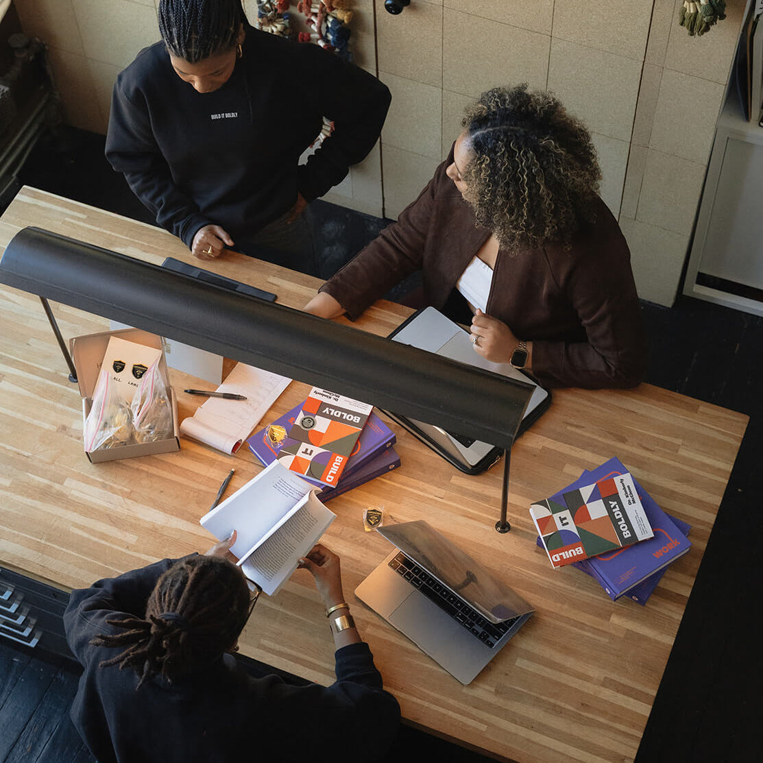 Overhead view of a creative team meeting around a wooden table with laptops, notebooks, and copies of Build It Boldly and The Leader’s Work.