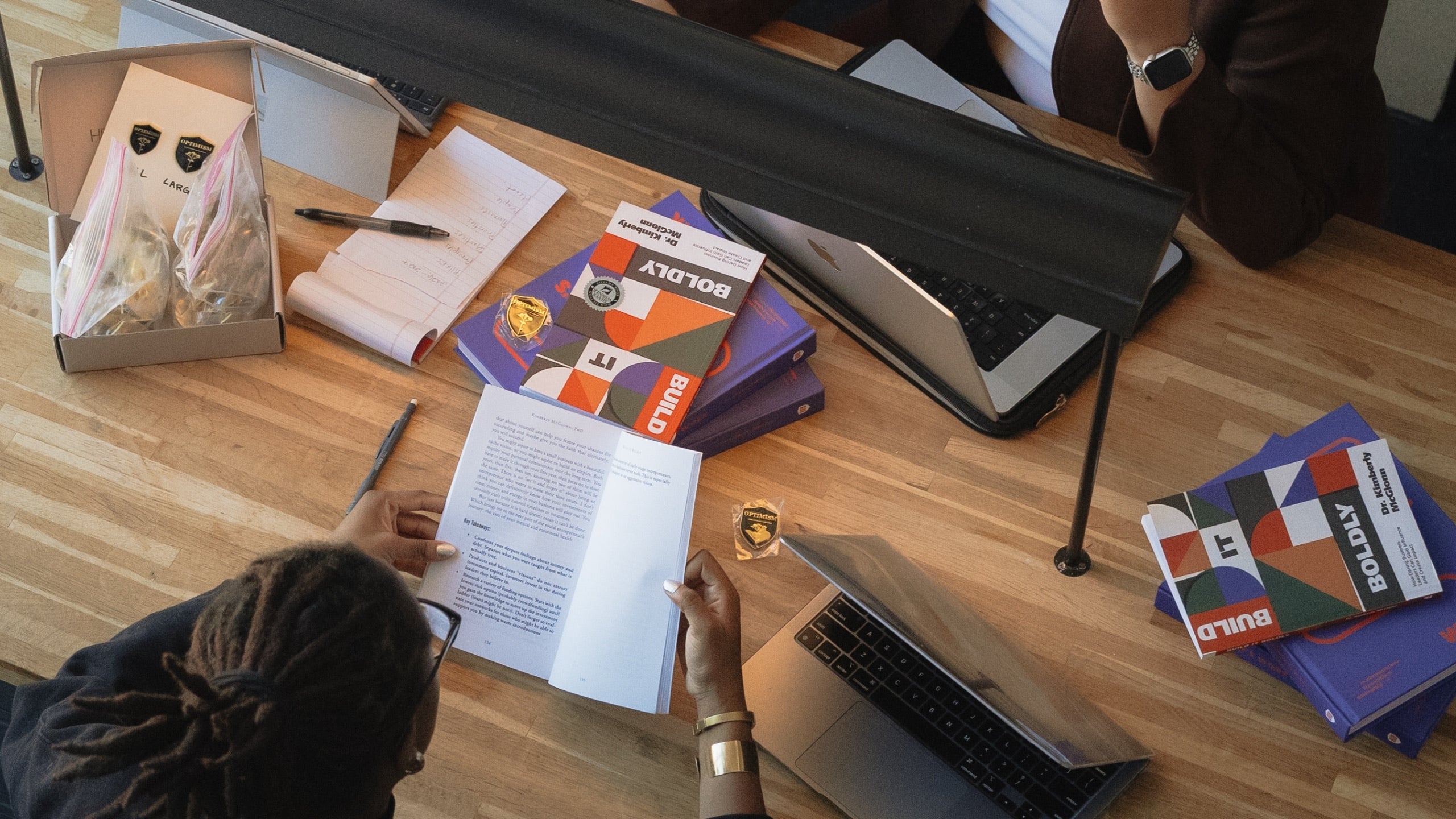 Overhead view of a Build It Boldly team workspace with laptops, notebooks, pins, Build It Boldly books, and The Leader’s Work workbooks on a wooden table.
