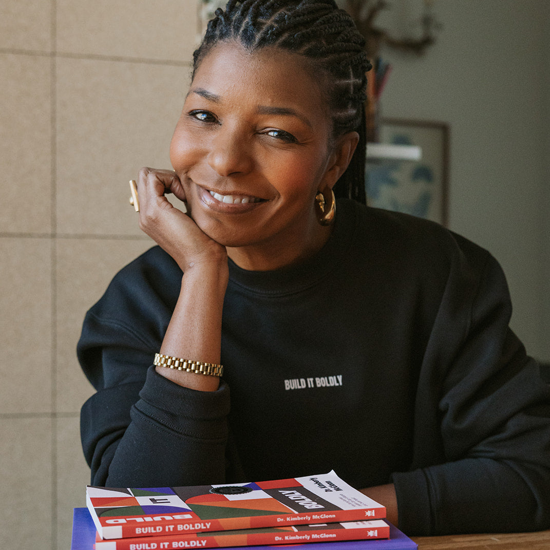 Portrait of Kimberly McGlonn smiling, wearing a black Build It Boldly sweatshirt, seated at a wooden table with stacked copies of Build It Boldly and The Leader’s Work by Dr. Kimberly McGlonn.