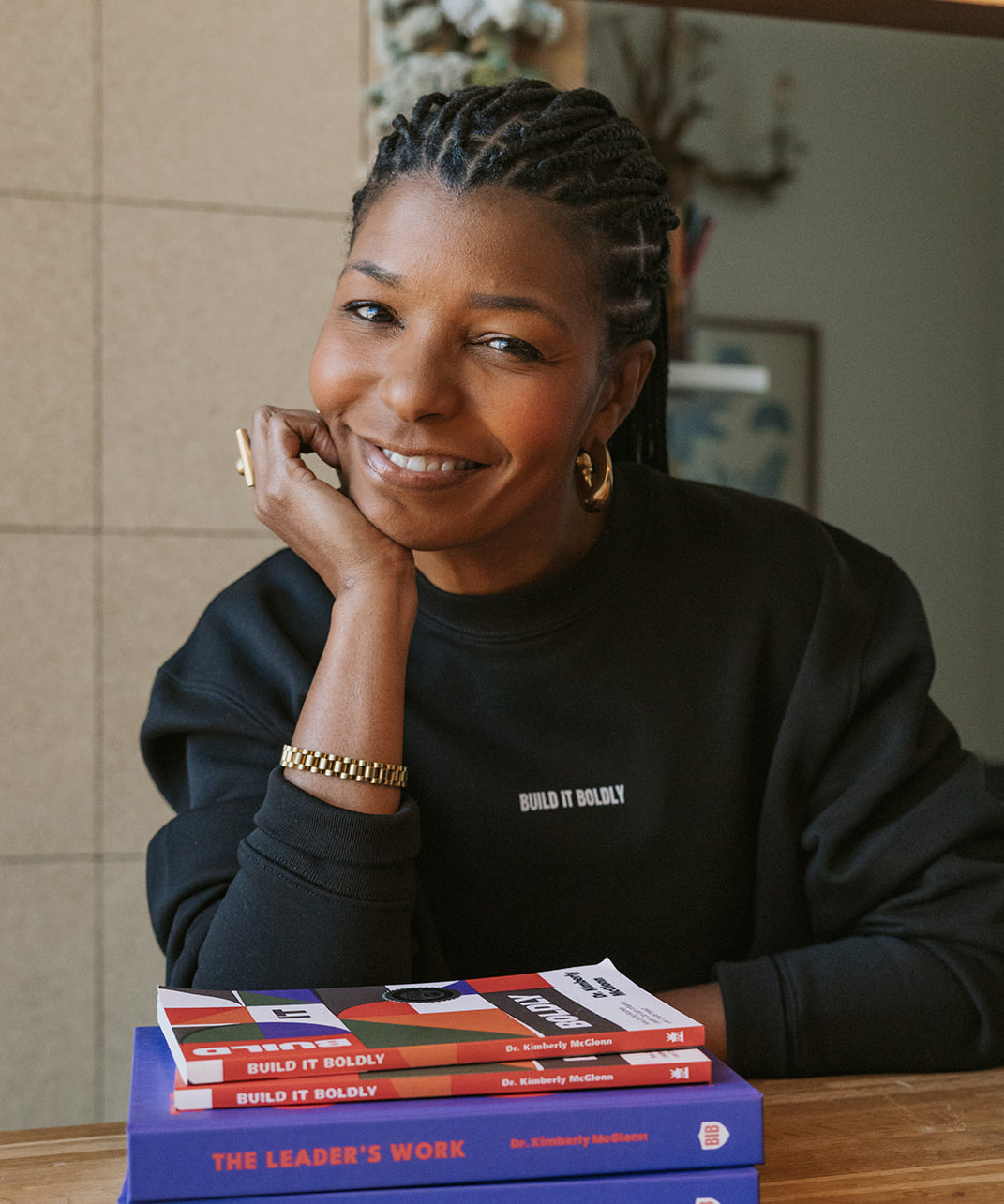 Portrait of Kimberly McGlonn smiling, wearing a black Build It Boldly sweatshirt, seated at a wooden table with stacked copies of Build It Boldly and The Leader’s Work by Dr. Kimberly McGlonn.