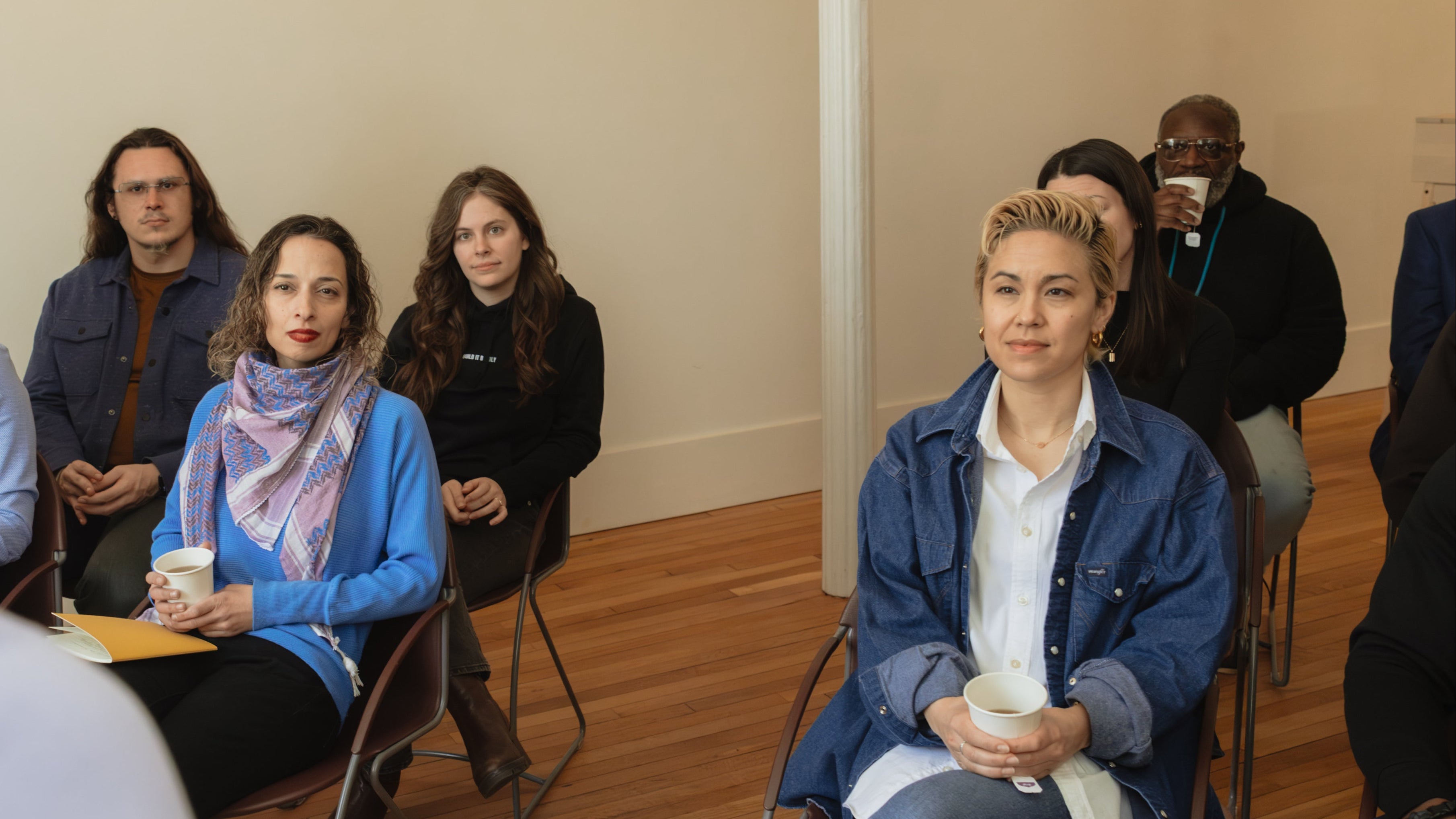 Audience members seated in a workshop or leadership session, listening attentively while holding coffee cups and notebooks in a bright room.