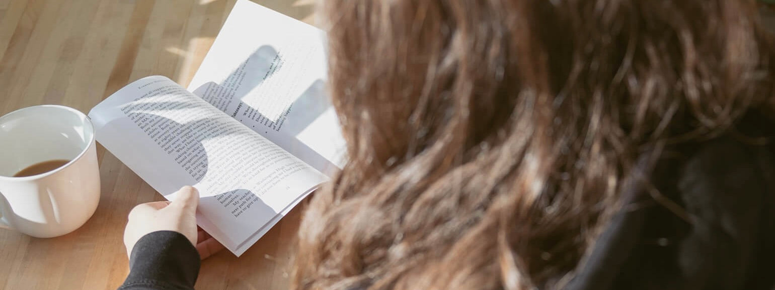 Person reading a book at a wooden table with coffee in natural light, reflecting on leadership and personal growth